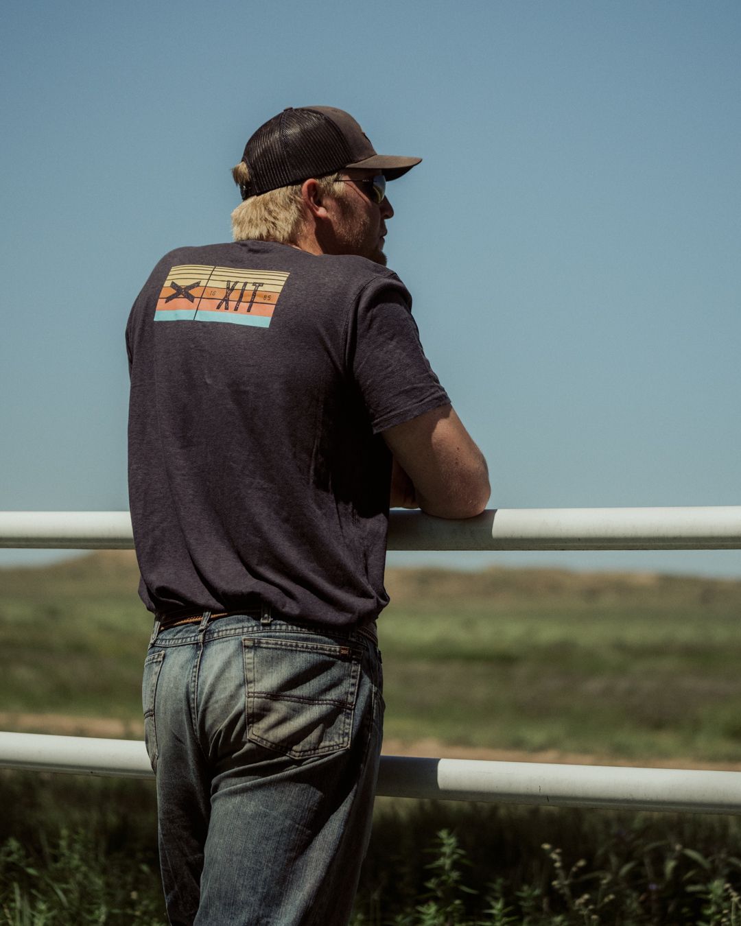 Photo of the back of a blonde male wearing a gray XIT t-shirt and a black and gray ball cap, leaning up against a fence