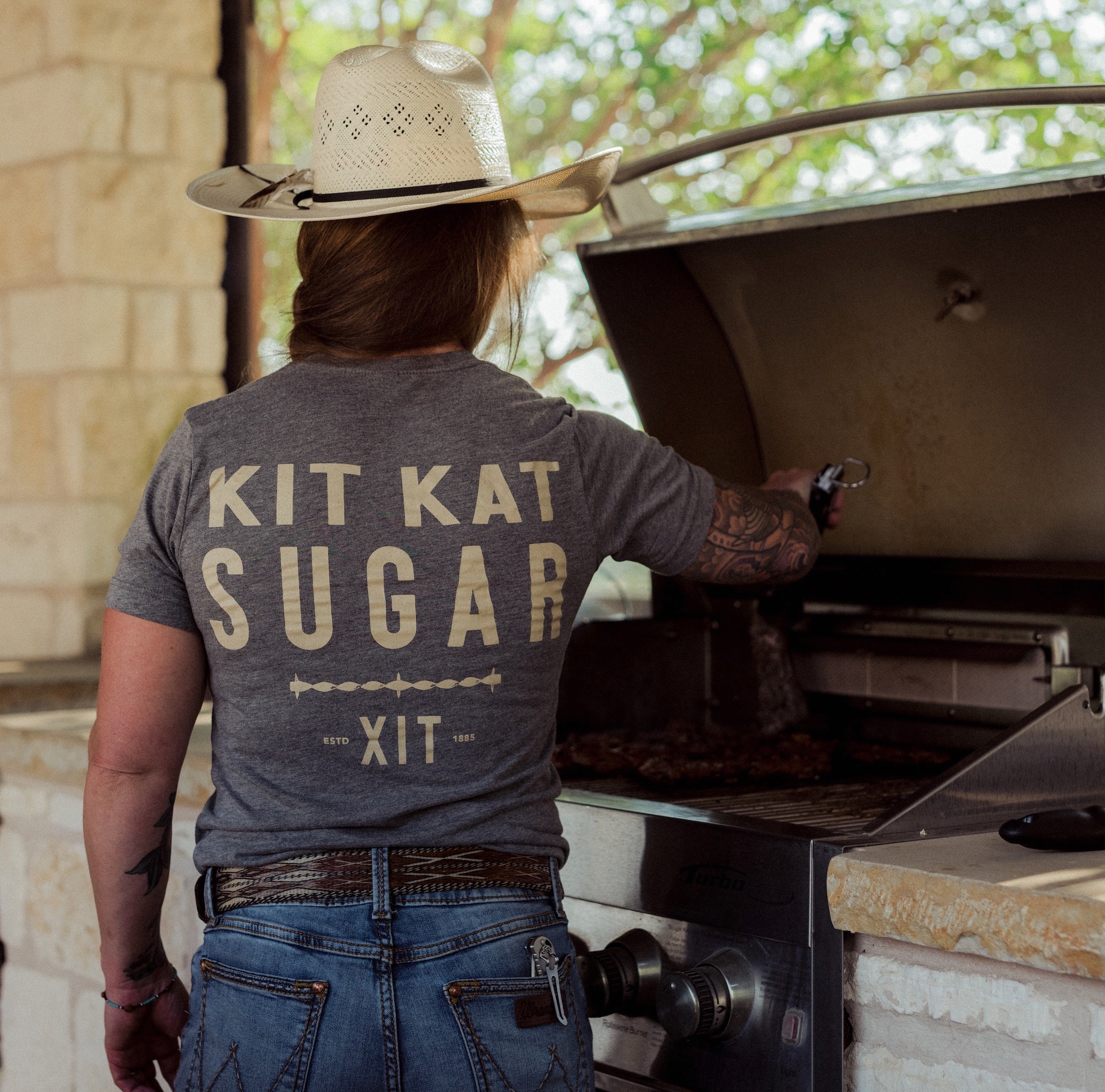 back of cowgirl grilling wearing gray t-shirt with Kit Kat Sugar and XIT text