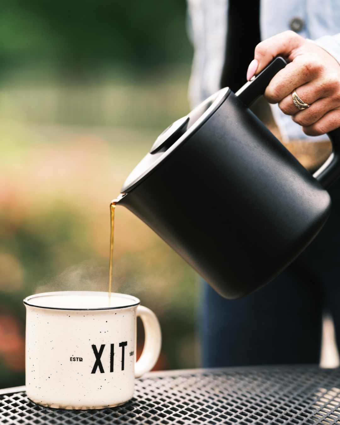 Person pouring coffee from a black kettle into a white mug with 'XIT' branding on a blurred outdoor background.