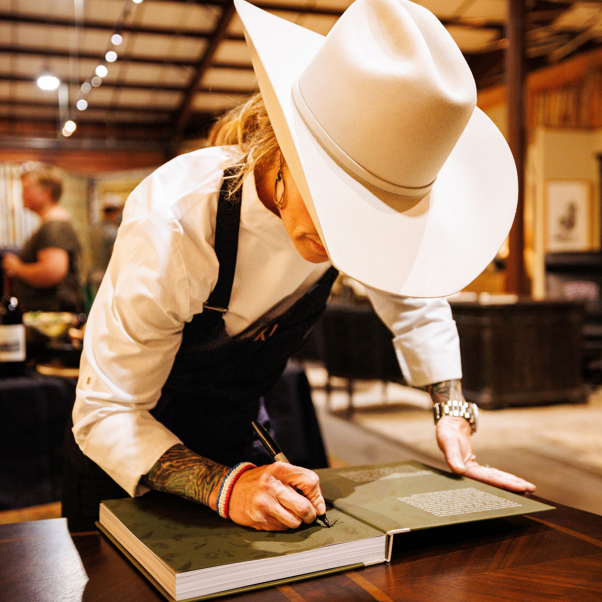 Cowgirl and Chef Abby Knowles wearing a cowboy hat and apron, signing The Modern Huntsman Cookbook: Recipes and Stories Earned in Wild Places cookbook on a wooden table with a black pen